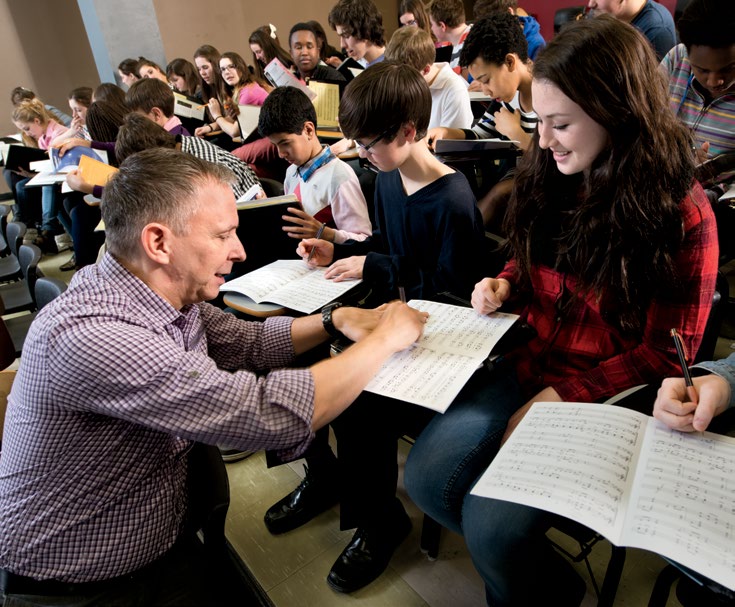 Photo de Robert Filion, dans une salle de classe, avec un grand nombre d’élèves. Il écrit dans la cahier de musique d’une élève.