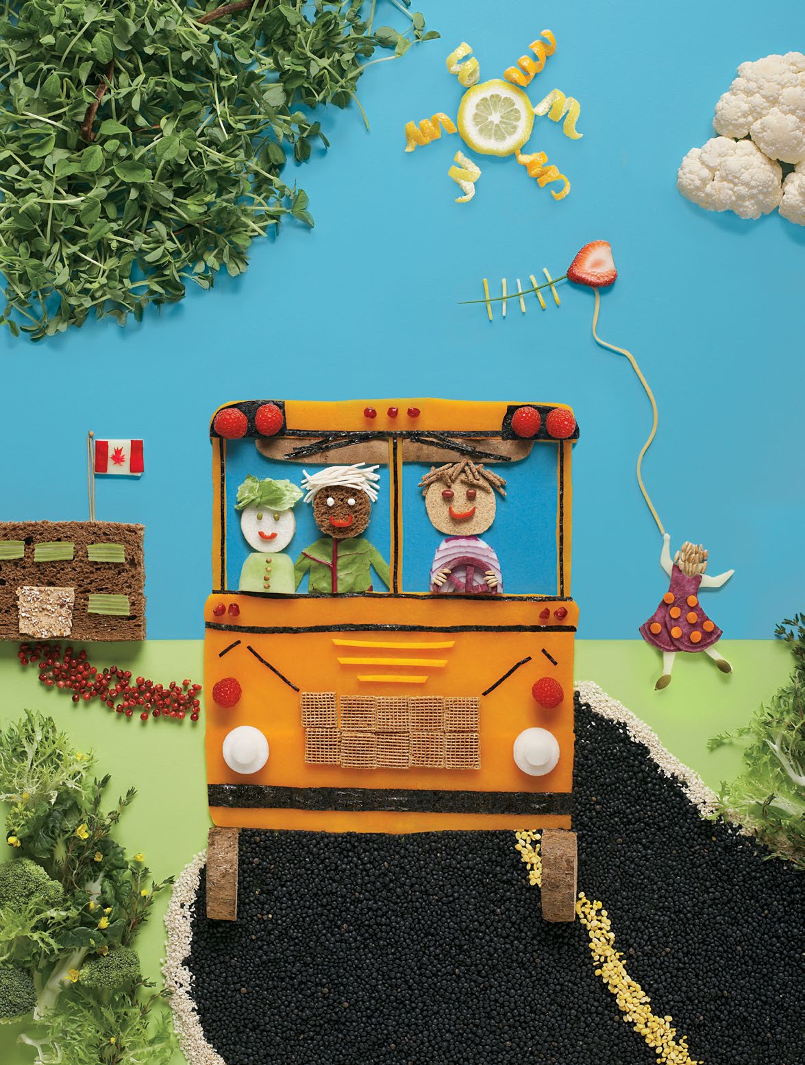 Illustration créée à partir de divers aliments. Un autobus scolaire est fait d’une tranche de courge et de framboises pour les phares. Un chauffeur et deux élèves sont à l’intérieur. Une tranche de citron représente le soleil; une fleurette de chou-fleur, des nuages; des feuilles de laitue, des buissons. Une élève lance un cerf-volant (tranche de fraise). Une tranche de pain, un craquelin et des tranches de céleri forment l’école où se dresse un drapeau du Canada.