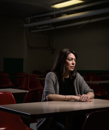 Photo d'une enseignante assise à un pupitre dans une salle de classe vide. La pièce est obscure, les mains de l’enseignante reposent l’une sur l’autre sur la table. Elle a un regard vide.