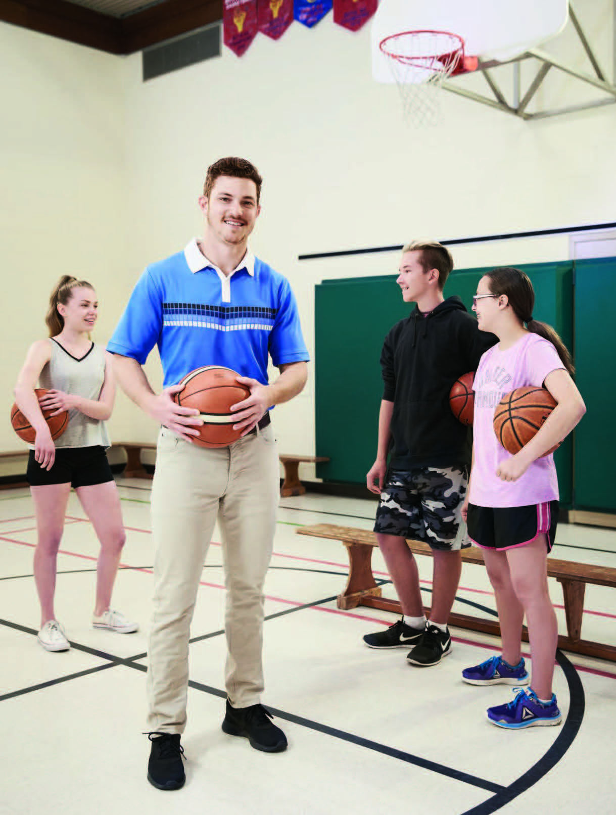 Photo de Corey Way, enseignant agréé de l'Ontario, debout dans un gymnase tenant un ballon de basketball dans les mains. Trois élèves sont derrière lui, chacun tenant un ballon de basketball sous le bras.