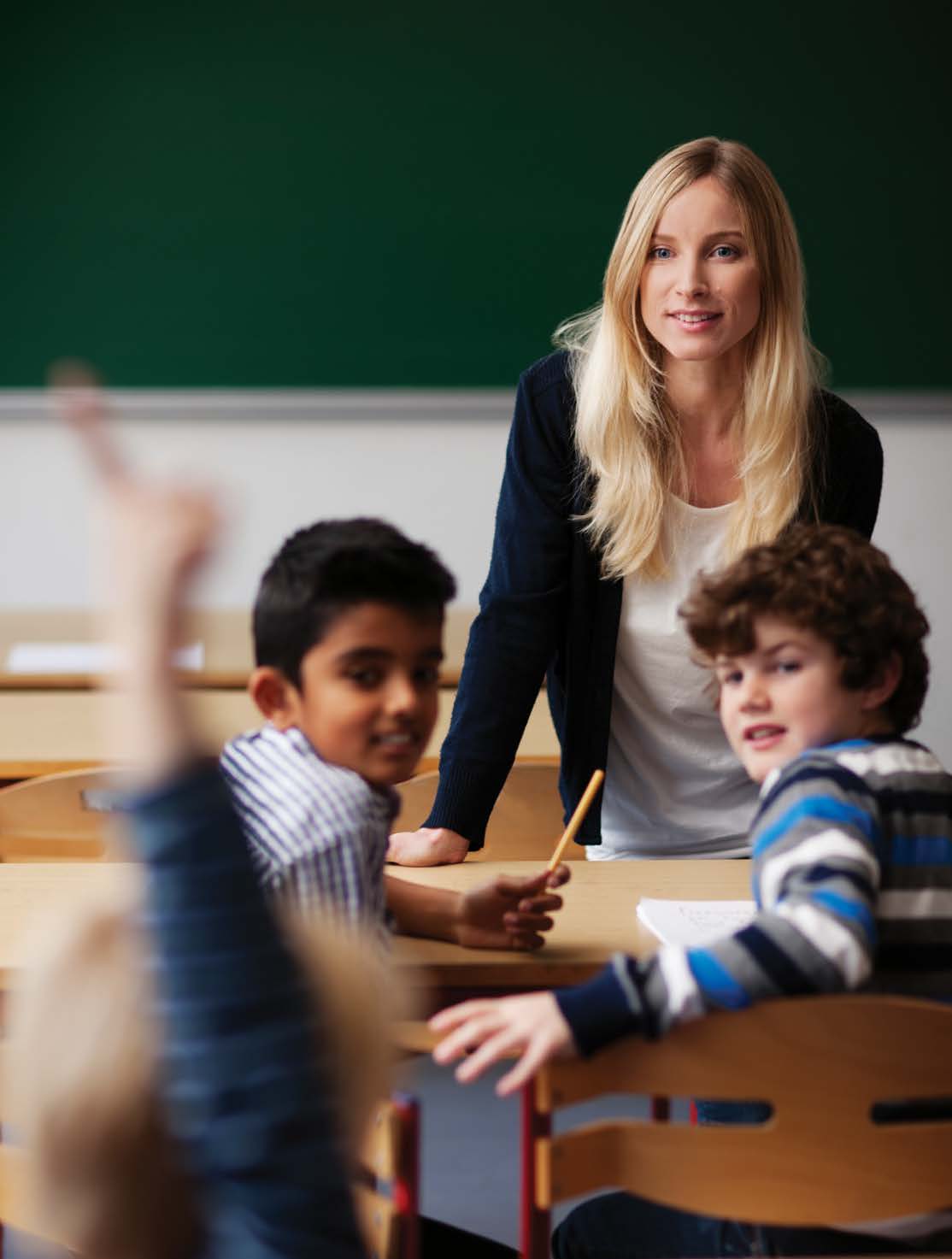 Photo d'une enseignante et de deux jeunes élèves qui regardent un élève qui a levé la main pour répondre à une question..
