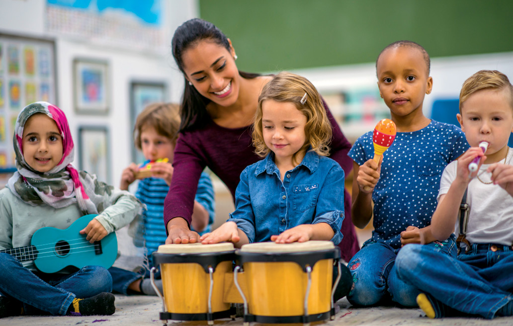 Photo d'une enseignante souriante ayant la main posée sur un tambour. Cinq jeunes élèves sont assis et jouent différents instruments de musique.