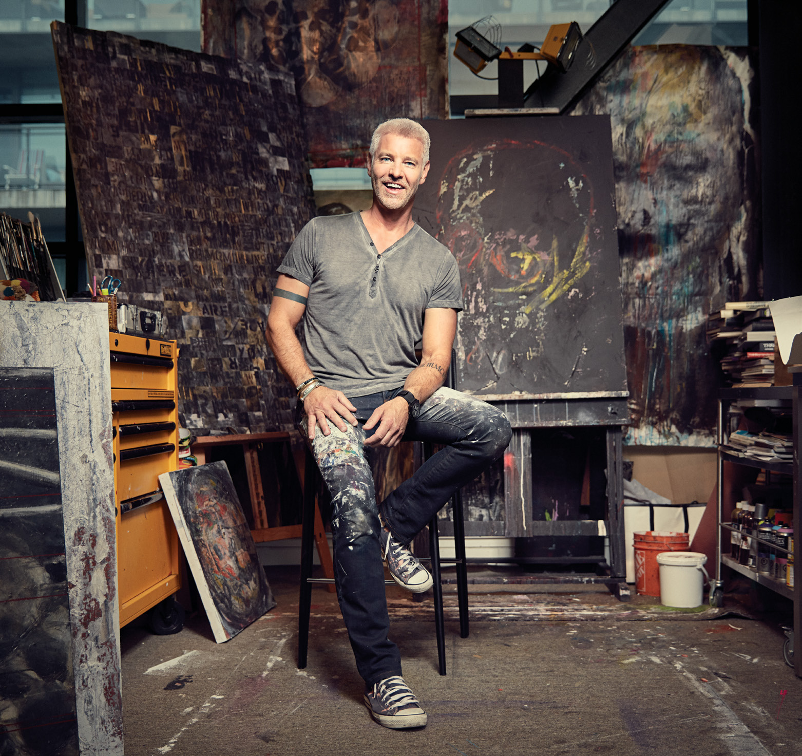 Photo de l'animateur de télévision et designer Steven Sabados assis sur un tabouret dans un studio avec, à l'arrière-plan, quelques œuvres peintes..
