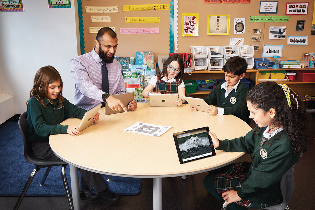 Photo d’Afzal Shaikh, enseignant agréé de l’Ontario, assis à une table avec un groupe de jeunes élèves regardant des tablettes électroniques.