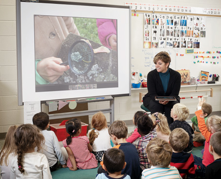 Photo d’Angela Harrison, enseignante agréée de l’Ontario, assise avec une tablette en main. Les jeunes élèves sont assis sur le plancher de la salle de classe. Ils regardent un grand écran.