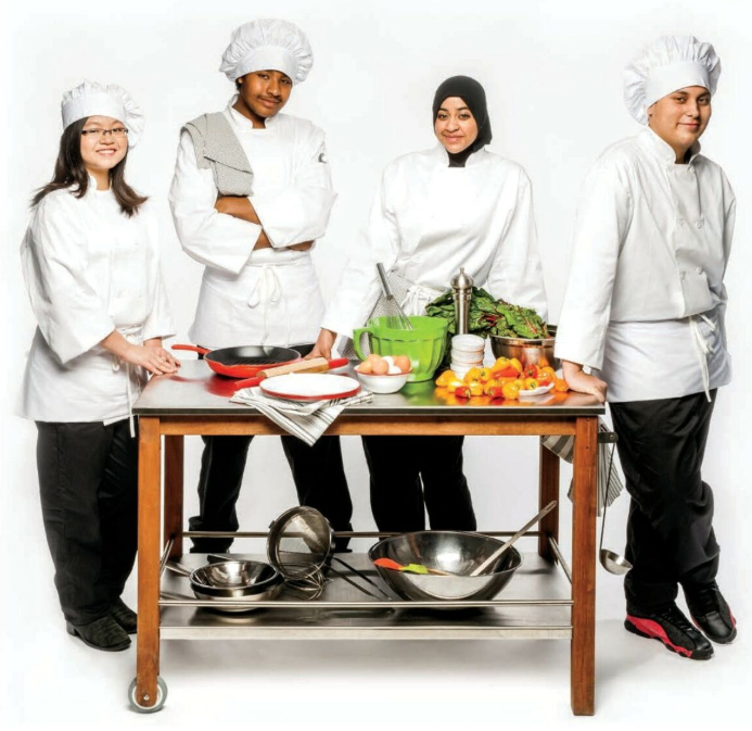 Photo de quatre élèves debout autour d’une table où se trouvent un poêlon, un rouleau à pâte, des œufs, des poivrons jaunes et rouges et des bols en acier inoxydable. Ils portent un pantalon, une veste et un chapeau de chef cuisinier.