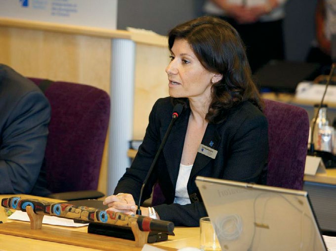 Photo de Liz Papadopoulos, présidente du conseil. Elle est assise à une table de réunion et parle au microphone.