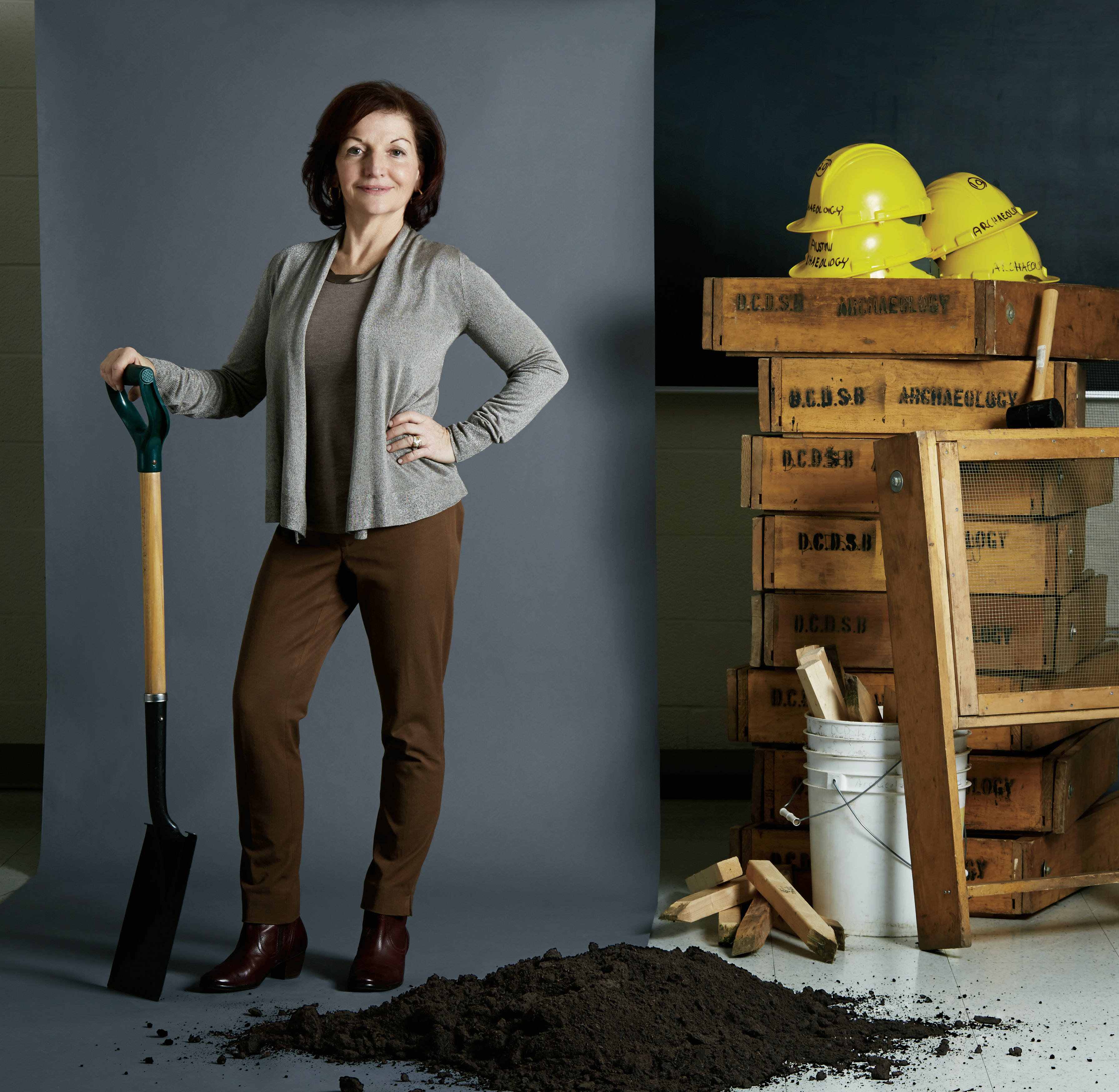 Photo de Catherine MacDonald, enseignante agréée de l’Ontario, debout, une pelle à la main. De la terre, de grands tamis, des casques de protection, un marteau et des chaudières sont empilés à sa droite.