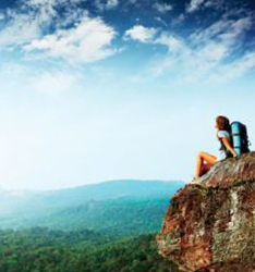 Photo d’une personne avec sac à dos. Elle est assise sur le bord d’un rocher et admire un panorama magnifique.