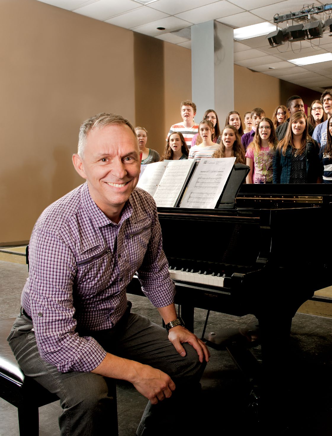 Photo de Robert Filion, enseignant agréé de l’Ontario, et musicien. Il est assis, souriant, sur le banc de son piano. Derrière lui, de nombreux élèves chantent en chœur.