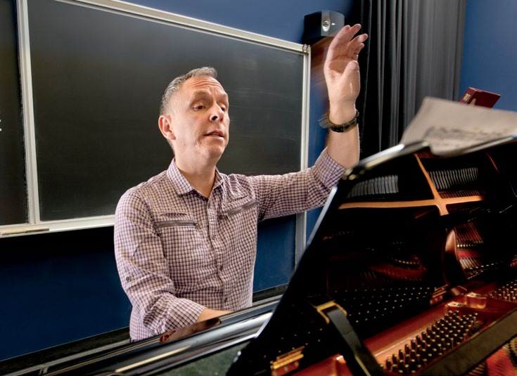 Photo de Robert Filion, au piano, la main gauche levée pour indiquer la bonne mesure à ses élèves. Un tableau noir, propre, est derrière lui.