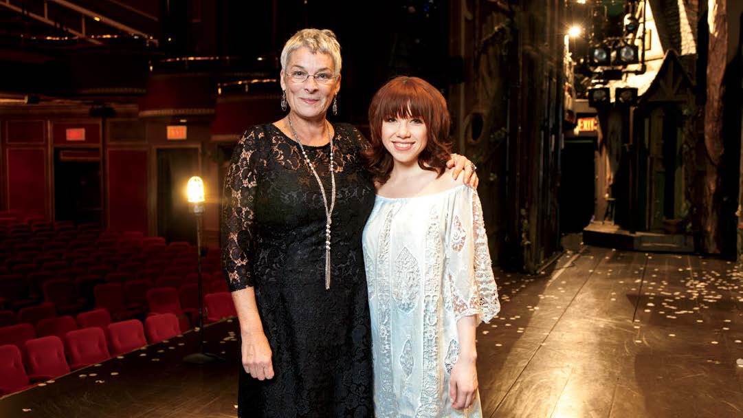 Photo de Beverley Holmes, enseignante de théâtre à la retraite, en compagnie de Carly Rae Jepsen sur la scène d’un théâtre. Les coulisses et les fauteuils en velours rouge créent le fond de scène.