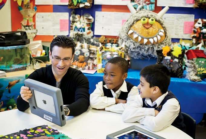 Photo de Vince Racanelli, enseignant agréé de l’Ontario, qui montre les applis d’une tablette à deux élèves. Un aquarium, des monstres comiques, des tableaux décorent la salle de classe.