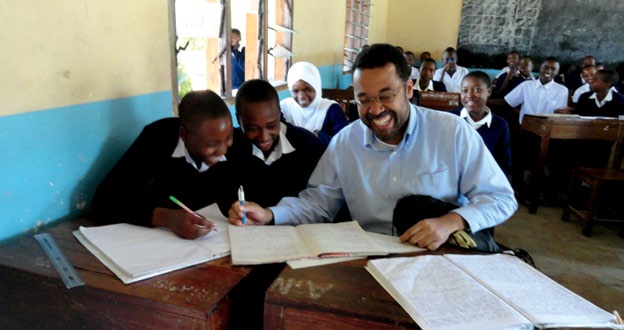 Photo d’une salle de classe en Tanzanie où l’enseignant est assis à un pupitre à côté de deux élèves.