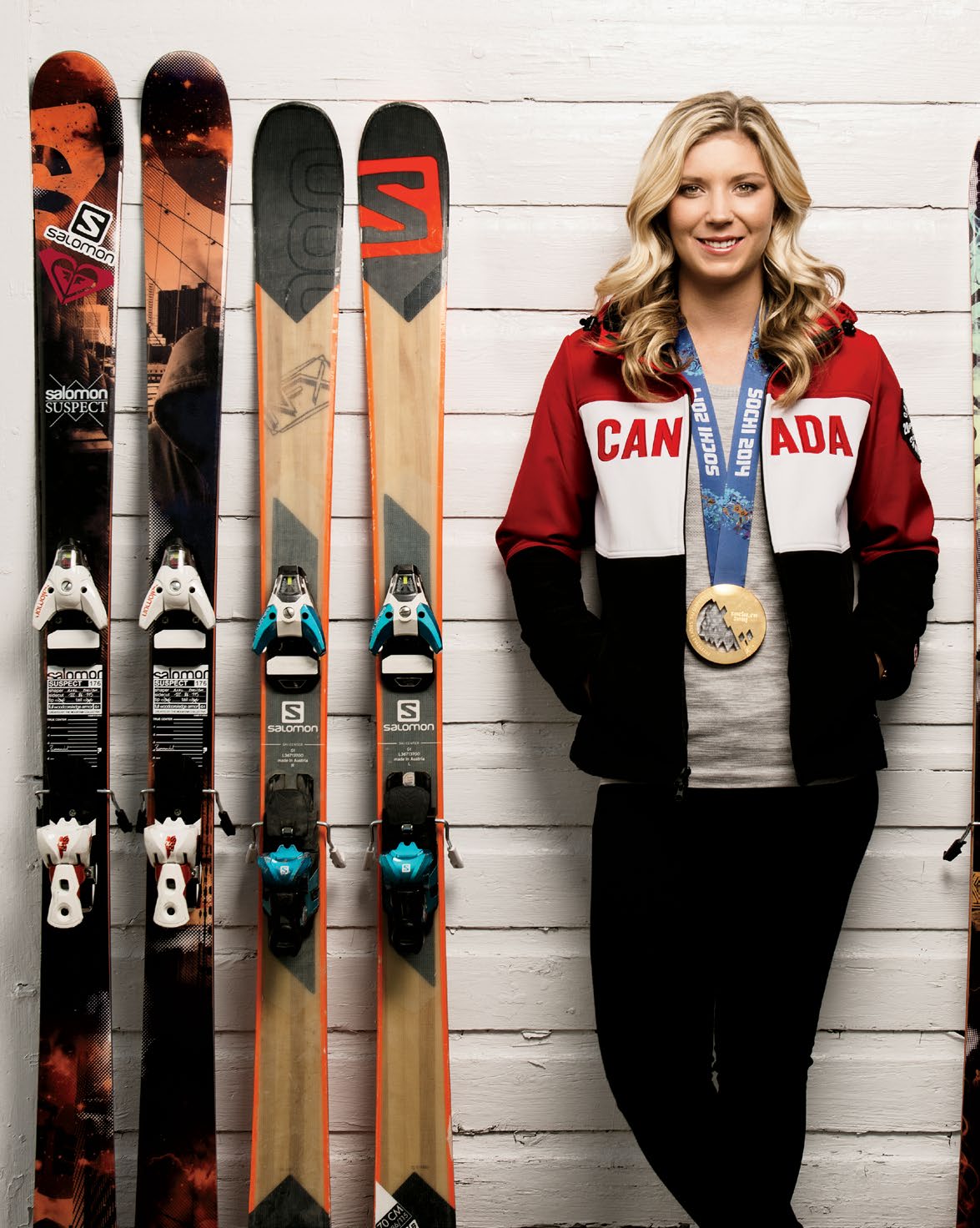 Photo de Dara Howell, skieuse olympique, appuyée contre le mur extérieur d’un chalet. Plusieurs paires de ski sont alignées, également contre le mur, de chaque côté d’elle. Elle porte sa médaille d’or olympique des jeux de Sotchi de 2014.