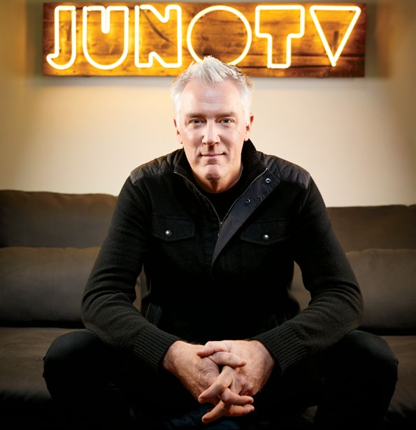 Photo d’Allan Reid, vétéran de l’industrie musicale, assis, les mains croisées. À l’arrière-plan, une enseigne en néon «Juno TV» est accrochée au mur.