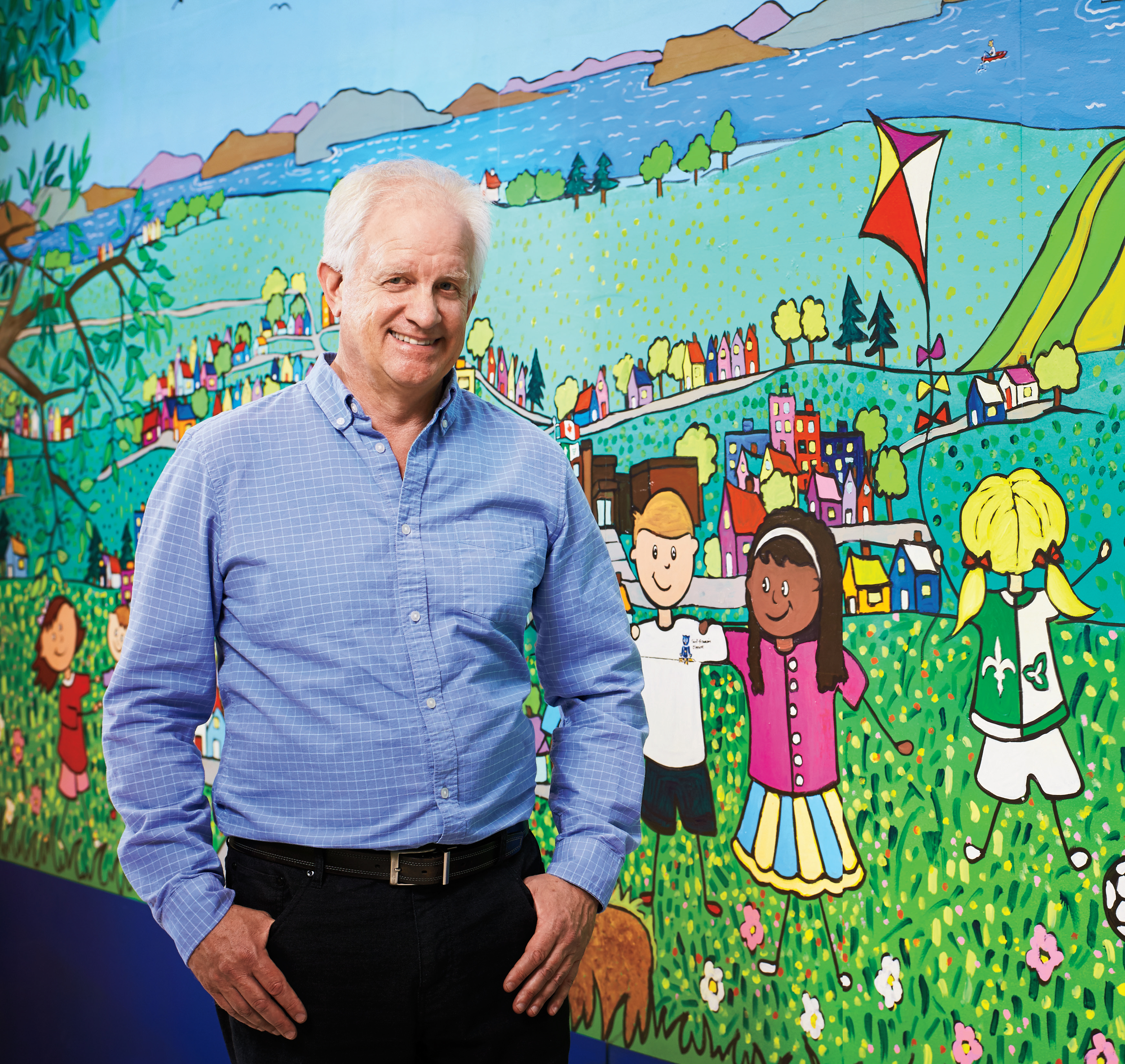 Photo d’André Roberge, enseignant agréé de l’Ontario, les mains dans les poches. Il est debout, devant une murale multicolore qui représente des enfants franco-ontariens, des maisons et des rues.