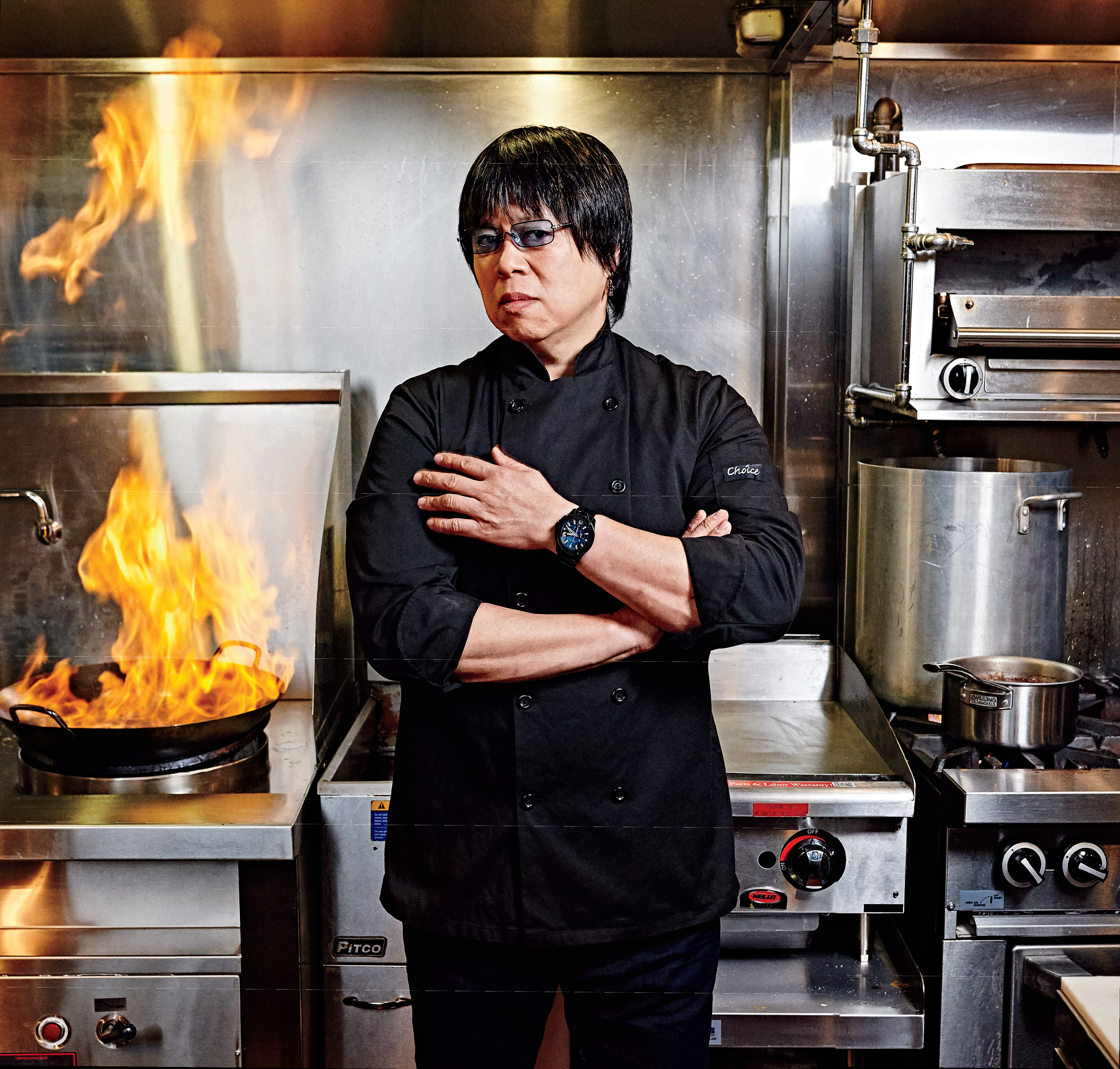 Photo d’Alvin Leung, juge de l’émission MasterChef Canada de CTV. Il se tient debout, les bras croisés, au milieu d’une cuisine commerciale. Des flammes s’élèvent d’un poêlon.