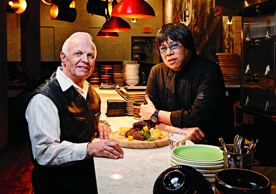 Photo du chef Alvin Leung en compagnie de Wayne Ellis, son ancien enseignant de 5e année. Ils sont debout, de chaque côté d’un comptoir, dans la cuisine du restaurant d’Alvin Leung.