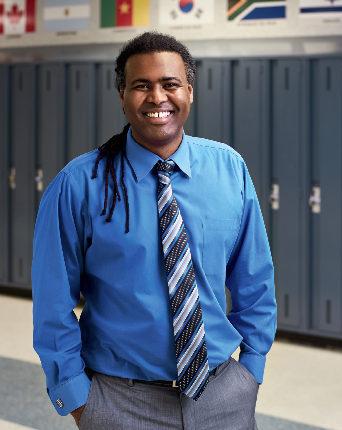 Photo d’Abdi Bileh Dirir, enseignant agréé de l’Ontario, debout, les mains dans les poches de son pantalon, dans un couloir de l’école où se trouvent les casiers des élèves.