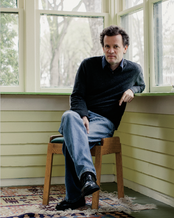 Photo d’Yann Martel, auteur à succès, assis sur un tabouret. Il est appuyé sur le rebord d’une fenêtre et regarde directement le photographe.