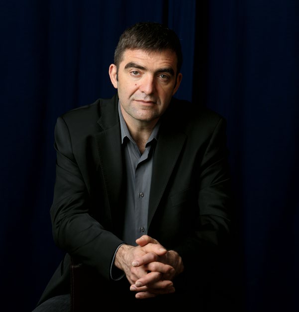 Portrait sur fond noir de Nino Ricci, auteur, assis avec les mains jointes.