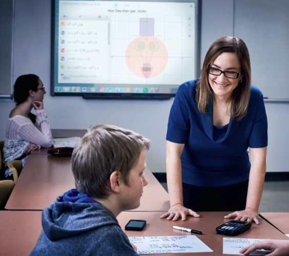 Photo de Martine Comeau, enseignante agréée de l’Ontario, debout en face de deux élèves, des garçons, assis à une table. À l’arrière-plan, une autre élève regarde le tableau.