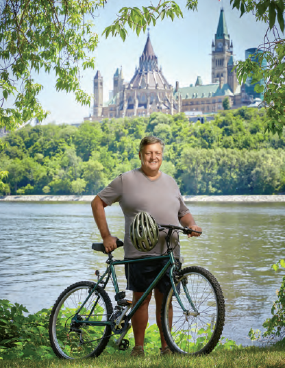 Photo de René Chiasson, enseignant agréé de l’Ontario à la retraite. Il est debout à côté de son vélo en face du canal Rideau. Les édifices du Parlement canadien figurent à l’arrière-plan.