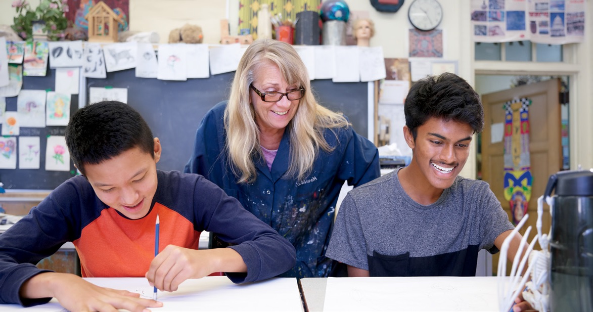 Photo d'une enseignante souriante regardant au-dessus de l'épaule de deux élèves qui travaillent sur un projet artistique.