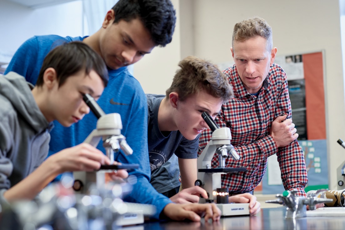 Photo d'un enseignant et de trois élèves. L'enseignant et un élève sont debout, tandis que les deux autres sont penchés en avant et regardent dans un microscope.