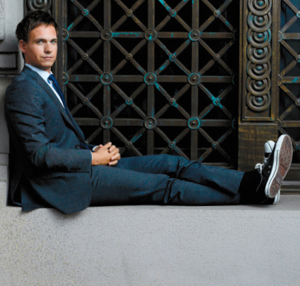 A photo of Suits star Patrick J. Adams sitting on a ledge with hands folded.