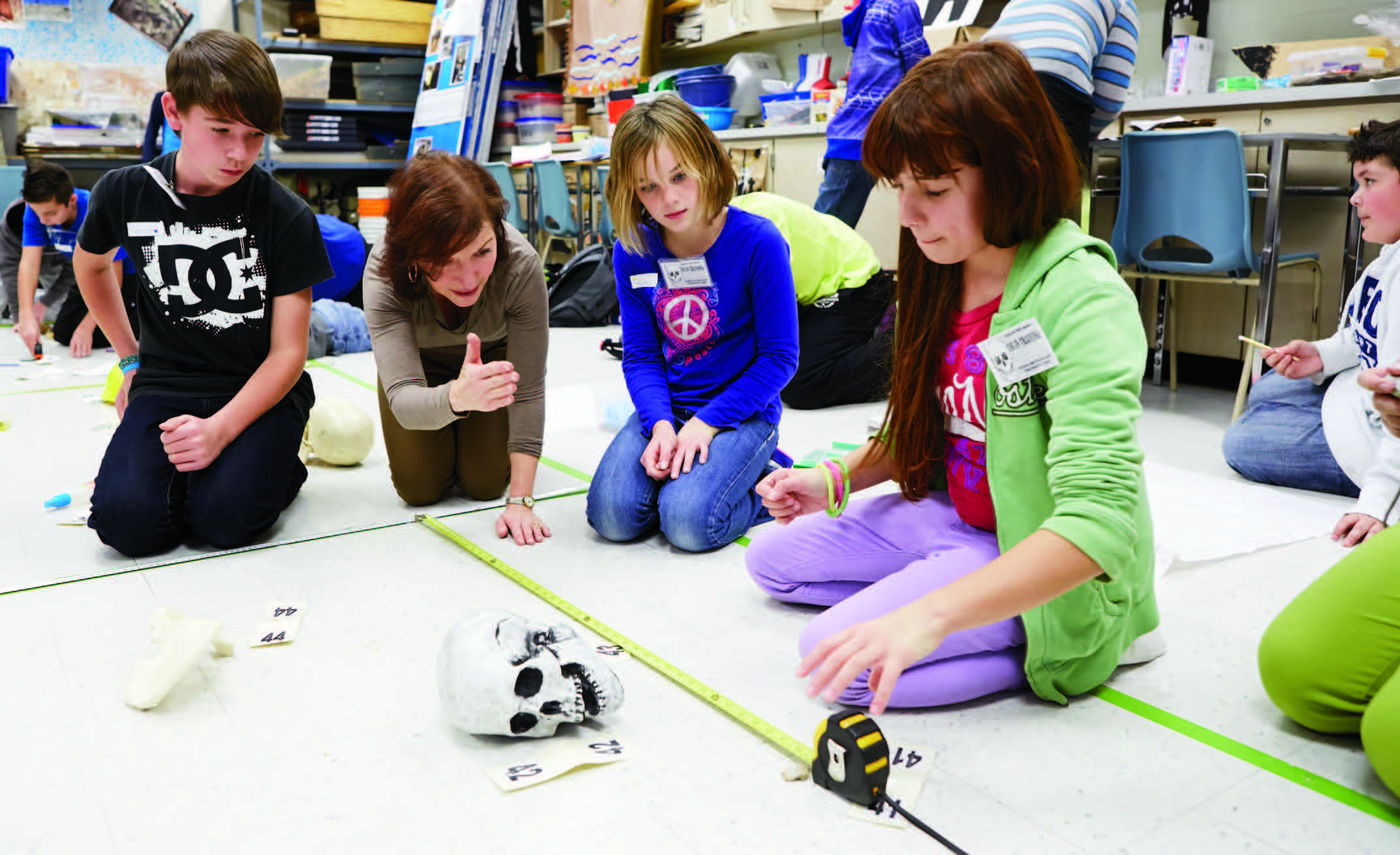 A photo of Catherine MacDonald and three young students kneeling behind a model human skull. Catherine is pointing at the scull and the students are measuring around it.
