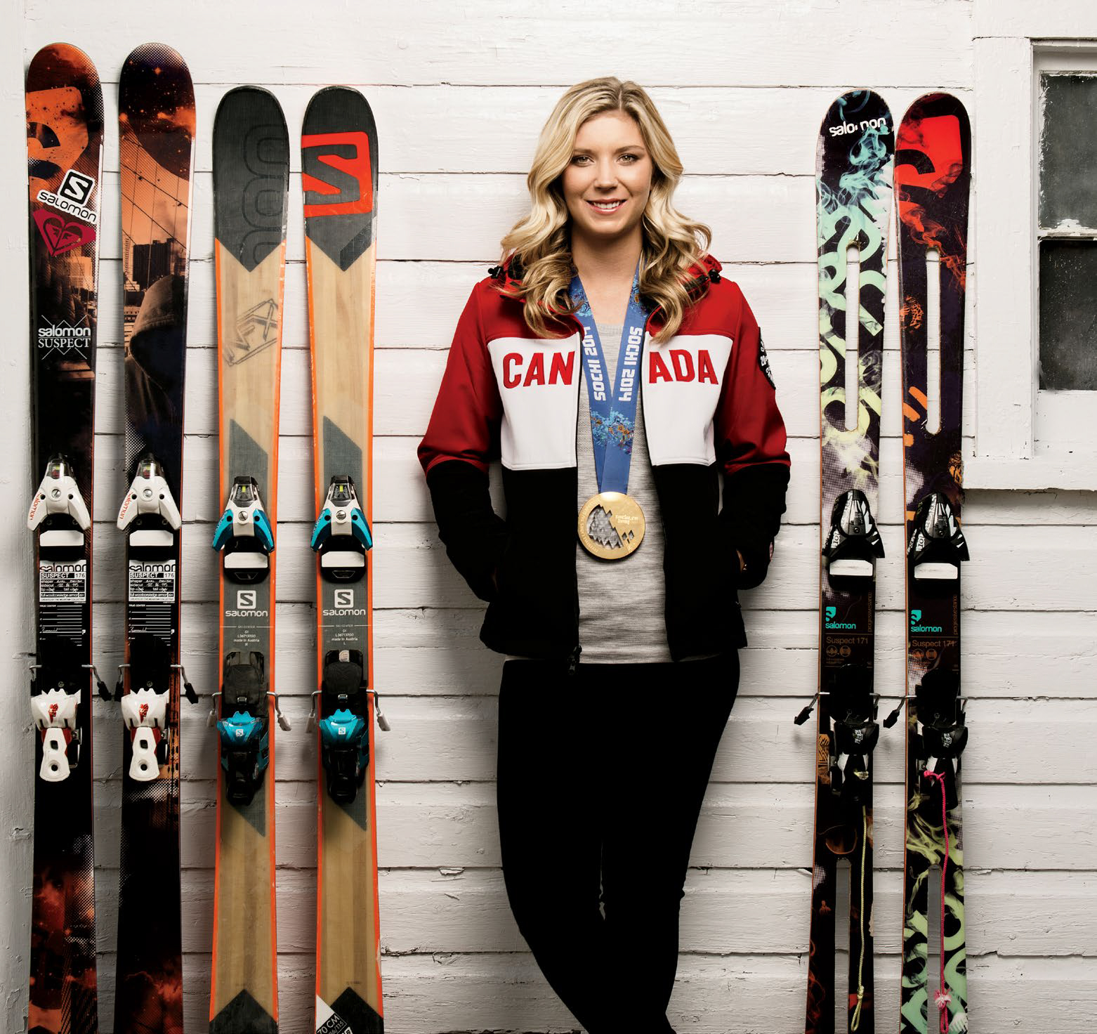 Olympic gold medallist Dara Howell standing with gold medal around neck. Dara is wearing a team canada jacket and flanked by skis.