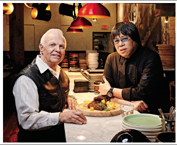 Photo of Alvin Leung with former Highland Heights Grade 5 teacher, Wayne Ellis. They are seated at a table in a kitchen with a cut of meat on the table.