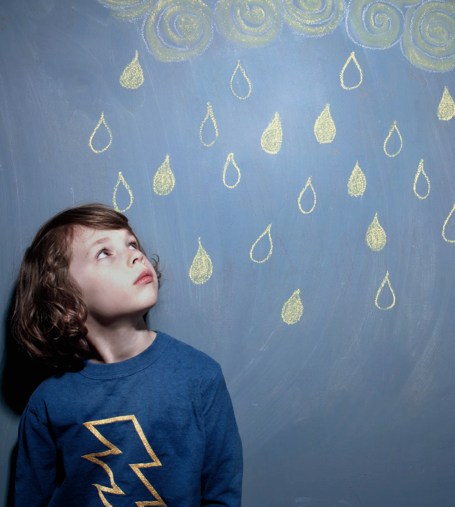A young student standing in front of a chalkboard and looking upwards. There are chalk raindrops above the student.