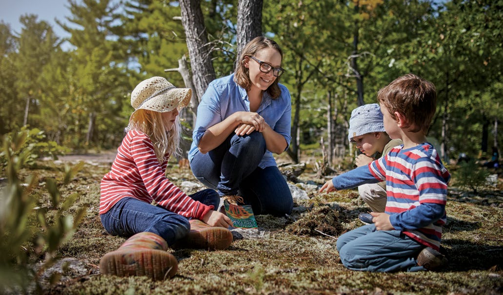Petra Eperjesi, crouching next to three young students in the forest. One of the students is pointing at the ground. Petra is smiling at the student.