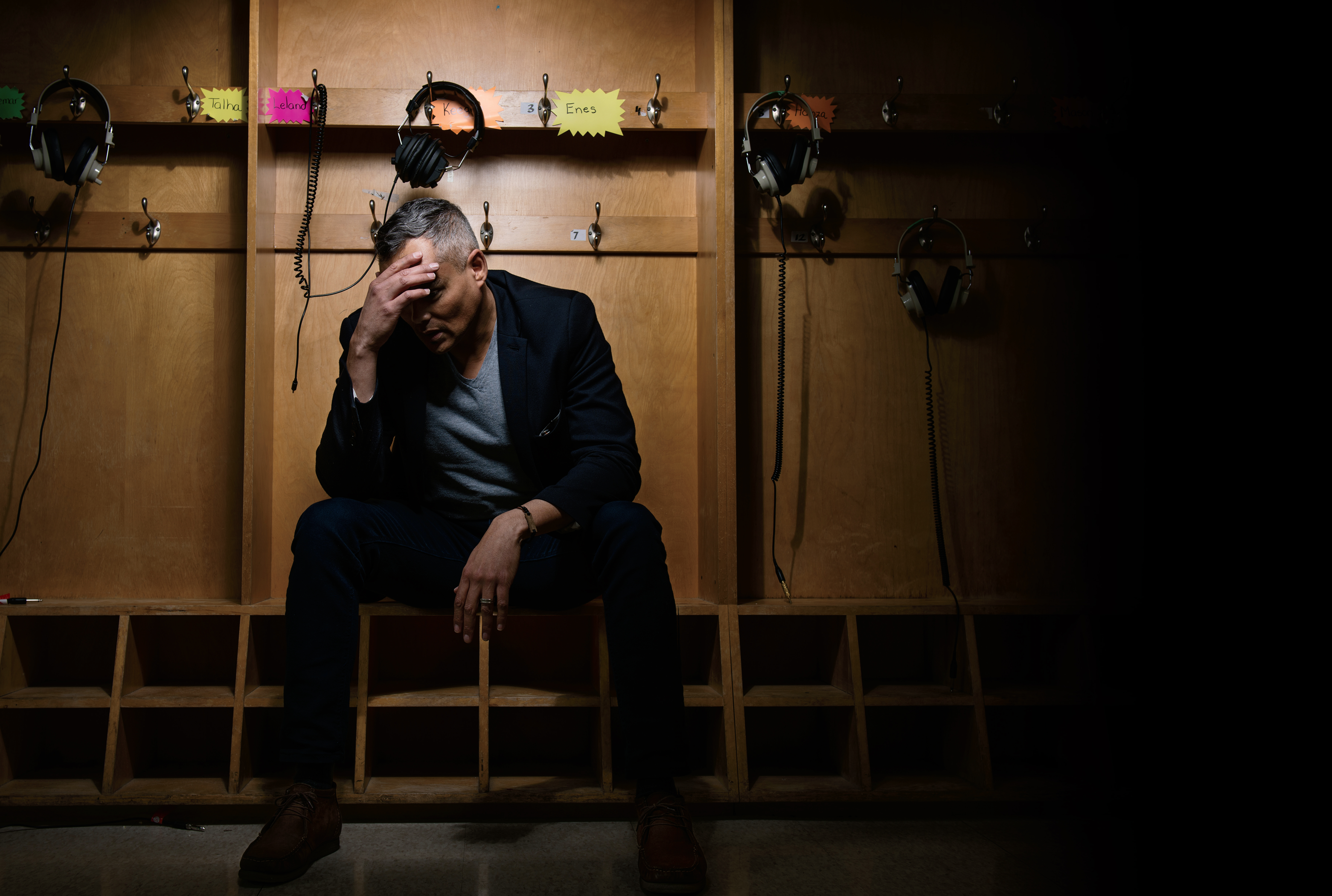 An image of a teacher in a locker room. The locker room is dark and the teacher is seated with a hand on their forehead while looking down. The teacher appears distressed.