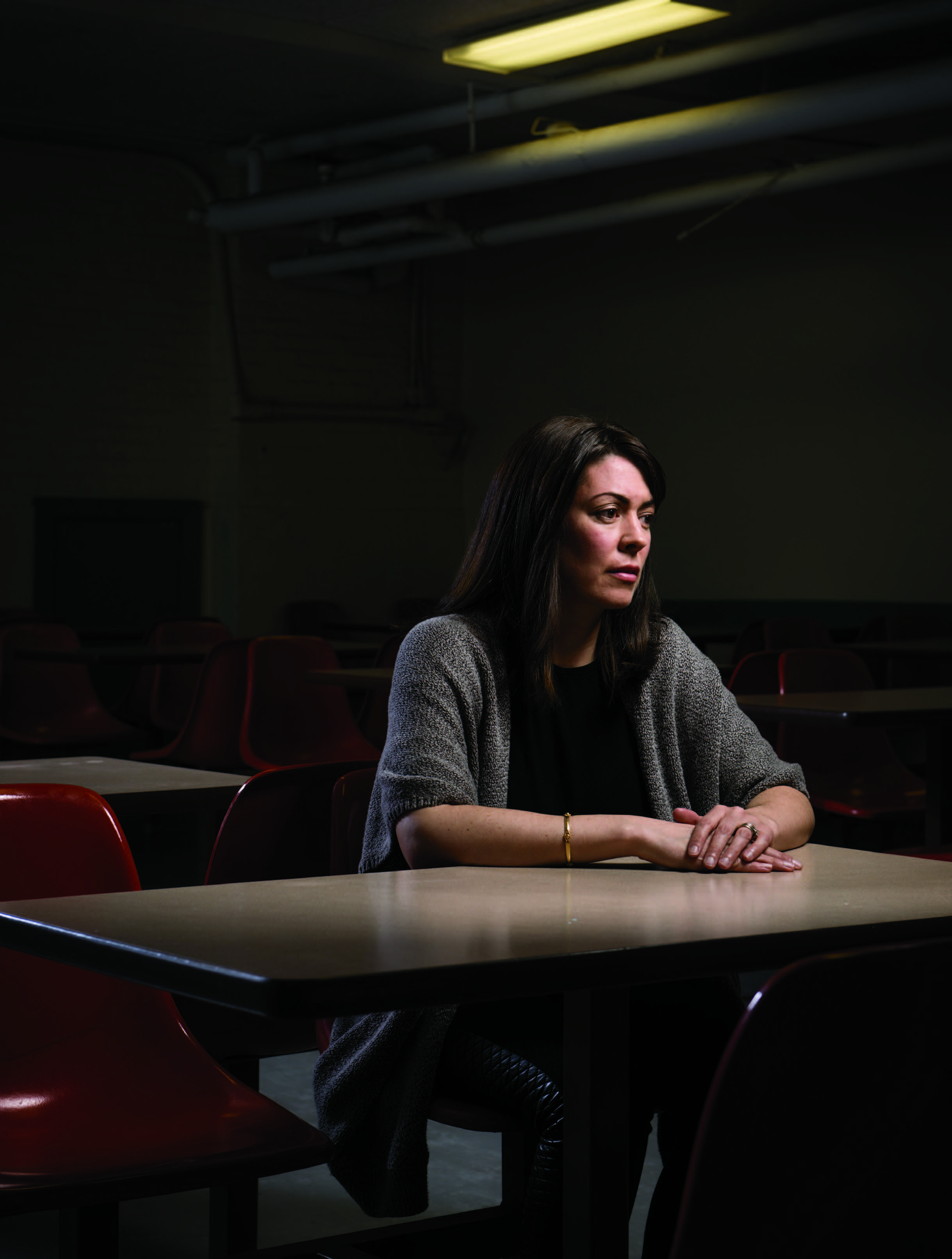 An image of a teacher seated at a desk in an empty classroom. The classroom is dark and the teacher has their hands folded on the desk. The teacher is staring blankly.