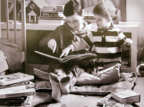 Black and white photo of a young student reading from an open book to a younger student. Both are seated on the floor surrounded by books. The younger student is pointing to the open book.