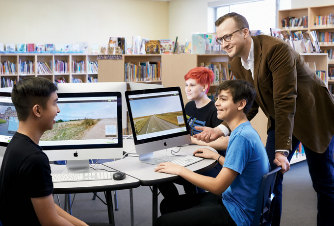 Photo of Ryan Henderson, Ontario Certified Teacher speaking with a group of young students. The students are seated at a table and working on computers.