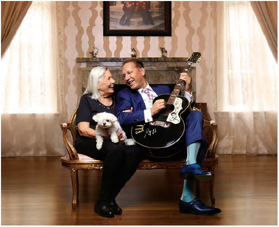 Photo of Dragons' Den star and entrepreneur Michael Wekerle seated on a chair with his mother Hermine, they are both looking at each other and smiling. Michael is holding a guitar and Hermine is holding a lap dog. 