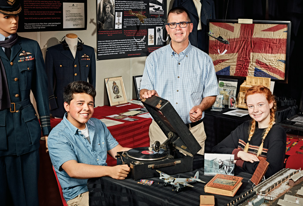A photo of Robert Flosman. Two young students are smiling and sitting at a table with a record player, airplane model, photograph and old book.