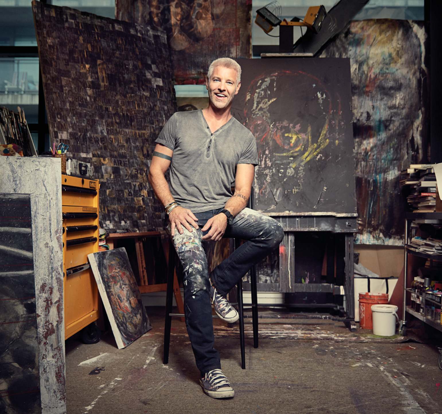 A photo of television host and designer Steven Sabados smiling and sitting on a stool in a studio in front of large paintings.