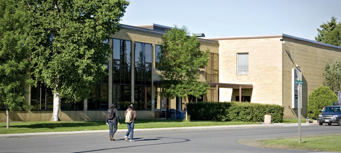 Photo of two students wearing backpacks walking along a road with a building and trees in the background.