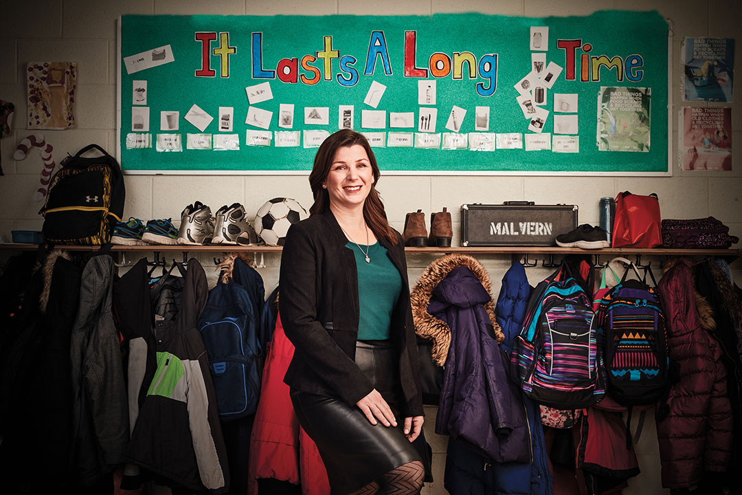 Photo of Rebecca Chahine, Ontario Certified Teacher, standing in front of a classroom setting with coats and backpacks hanging up.