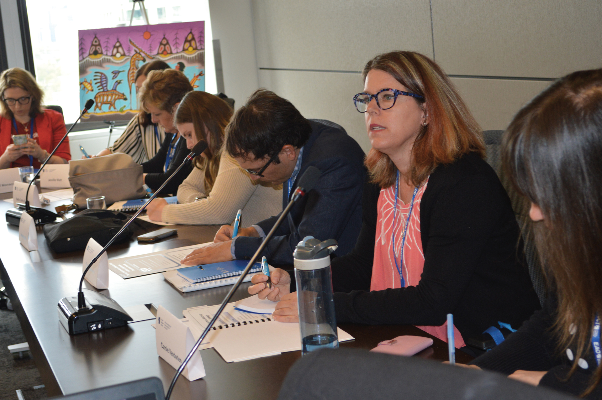 Photo of a person sitting down and speaking into a microphone during a meeting with several other people. 