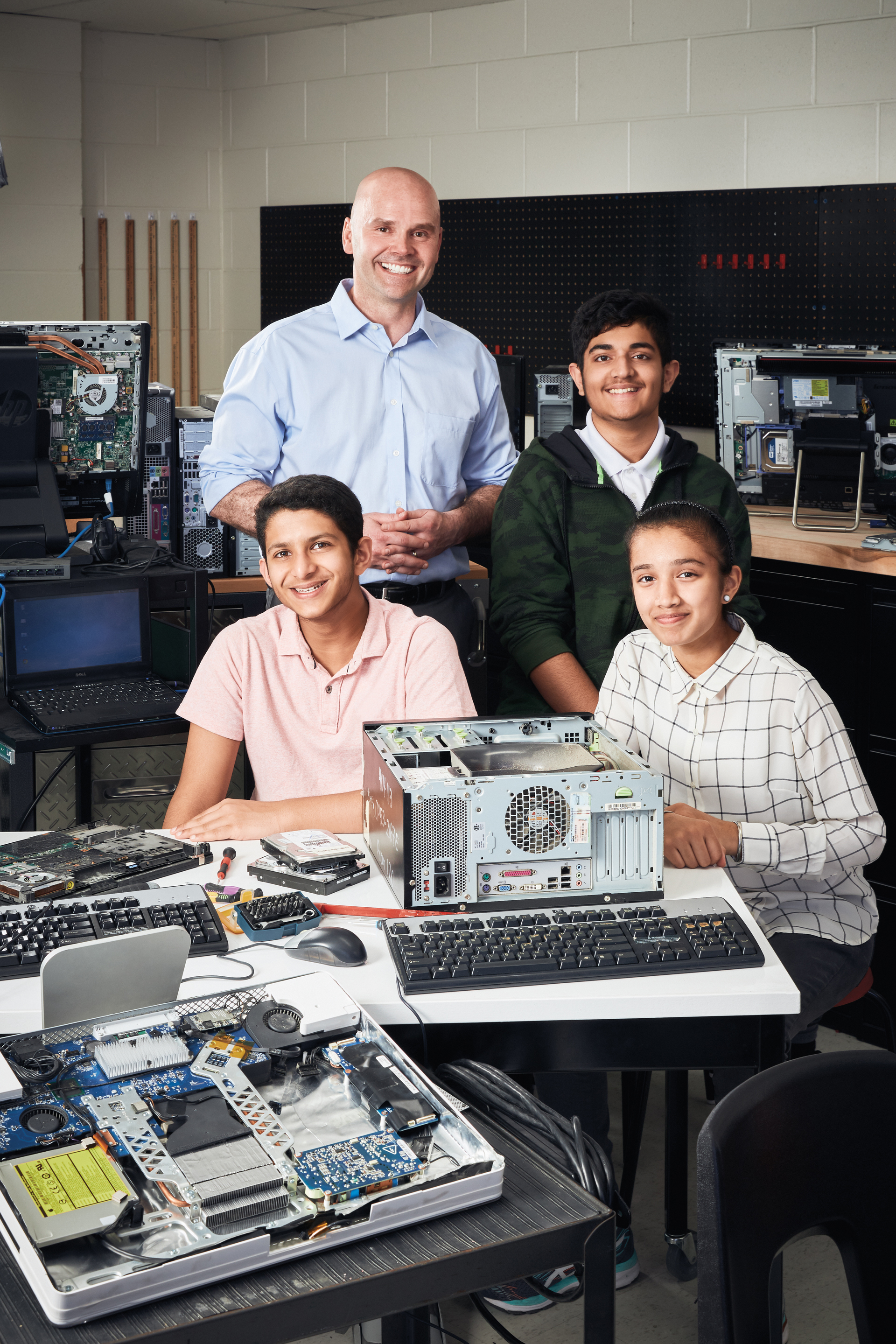 Photo of Andrew Dobbie, Ontario Certified Teacher, smiling with three smiling students in a computer classroom with several computer parts.