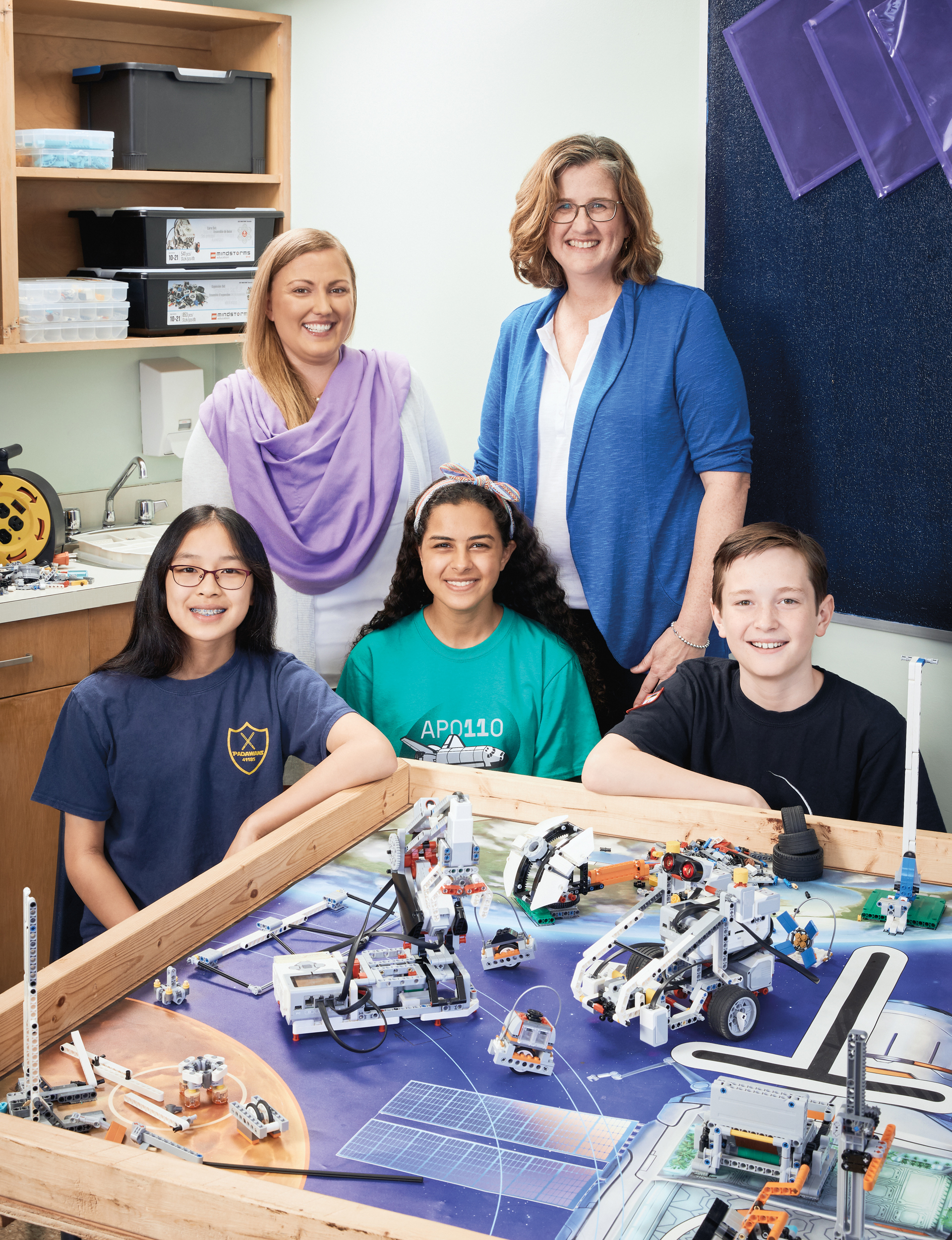 Photo of Shauna Nelson, Ontario Certified Teacher, and Sari McDowell, Ontario Certified Teacher, standing behind three students in a robotics classroom. In front of them are several robots made out of Lego.