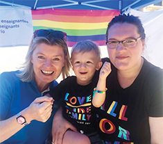 Photo of two people smiling with a smiling child in the middle. In the background is a flag with horizontal rainbow stripes.