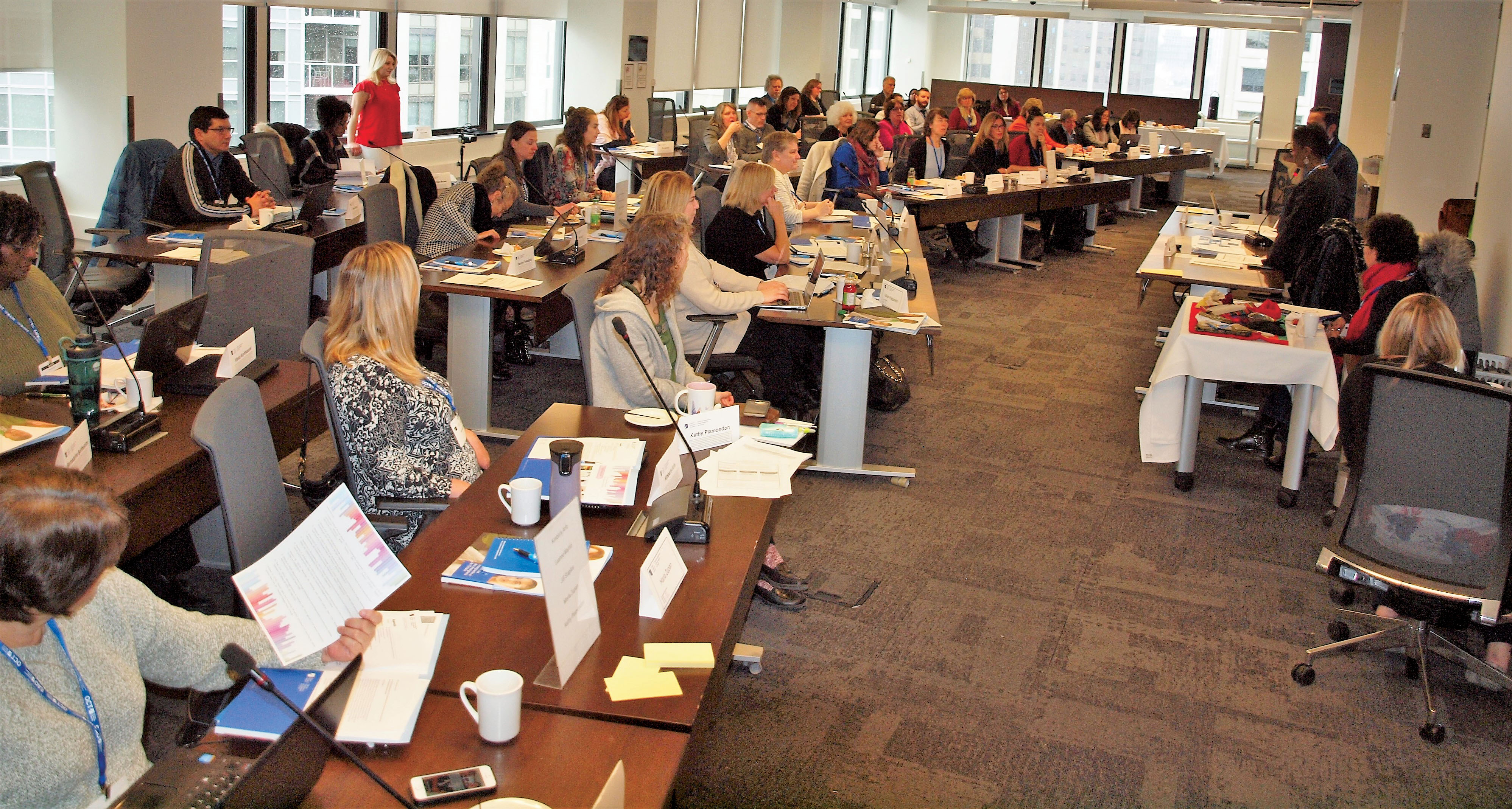Photo of a large group of people sitting at desks and facing a small group of people presenting.